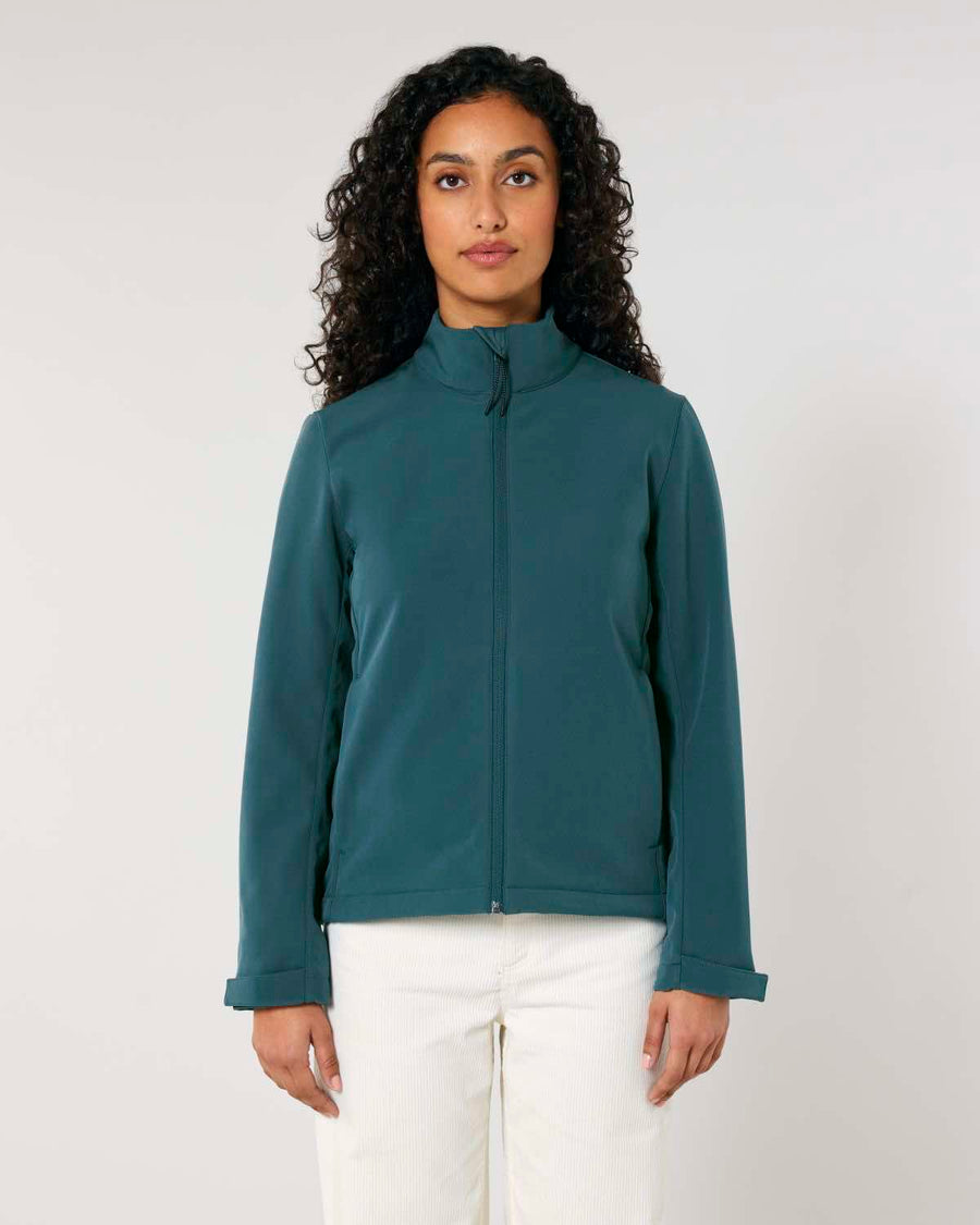 Woman standing against a plain background, wearing a MyNeedsAreSimple STJW166 Stanley/Stella Navigator Women’s Non-Hooded Softshell jacket in dark teal and white pants, looking at the camera.