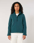 A woman with curly hair wearing a dark green MyNeedsAreSimple STJW166 Stanley/Stella Navigator Women’s Non-Hooded Softshell jacket and white pants, standing against a plain background.