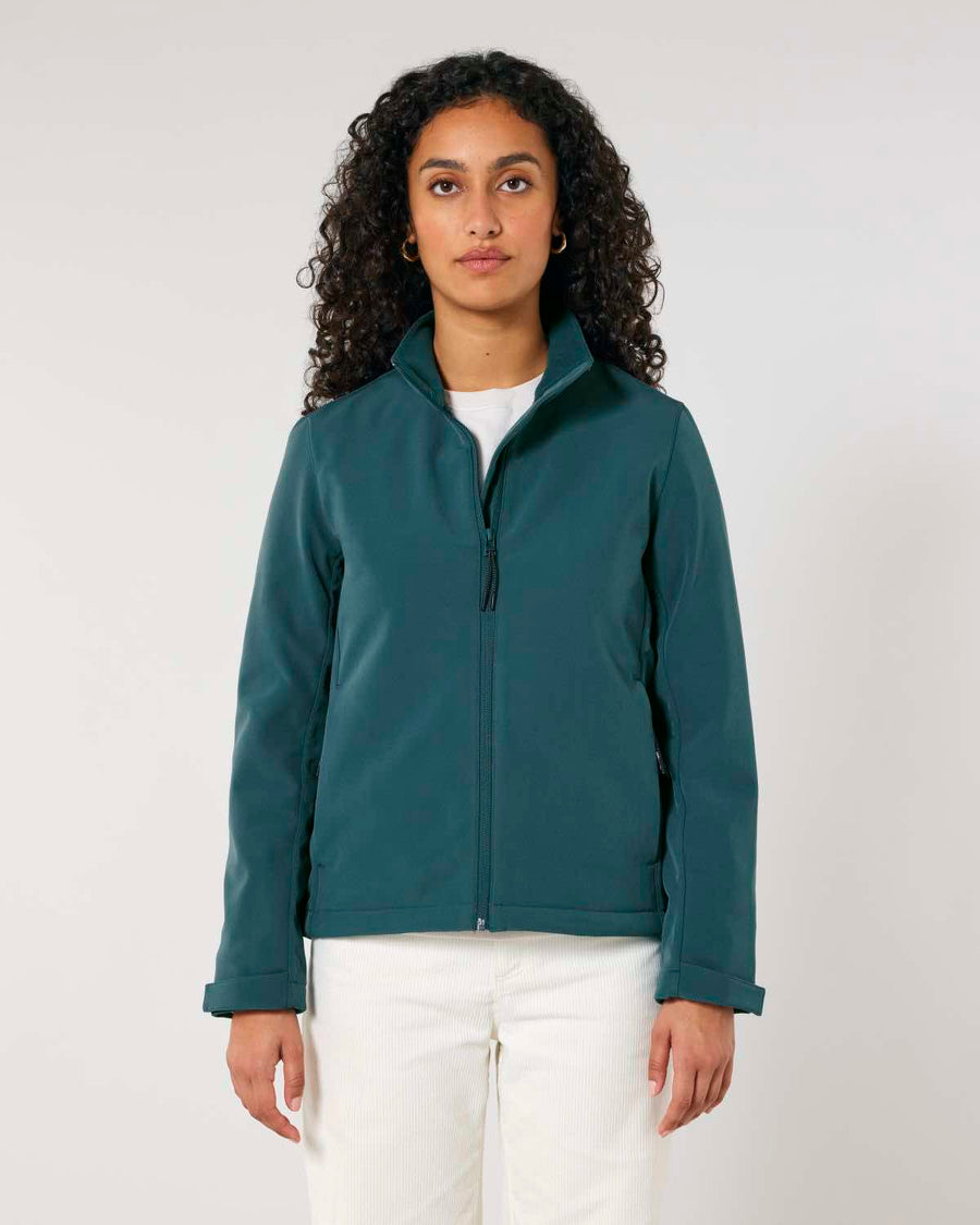 A woman with curly hair wearing a dark green MyNeedsAreSimple STJW166 Stanley/Stella Navigator Women’s Non-Hooded Softshell jacket and white pants, standing against a plain background.
