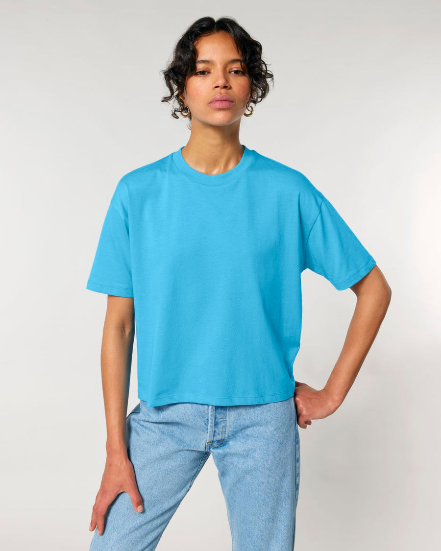 Woman posing in a light blue STTW175 Stella Nova Women's Boxy T-Shirt by Stanley/Stella and blue jeans against a neutral background.