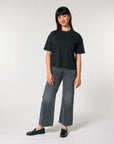 A woman standing against a neutral background, wearing a black Stanley/Stella Women's Boxy Fit T-Shirt made of organic ring-spun cotton, gray jeans, and black loafers.