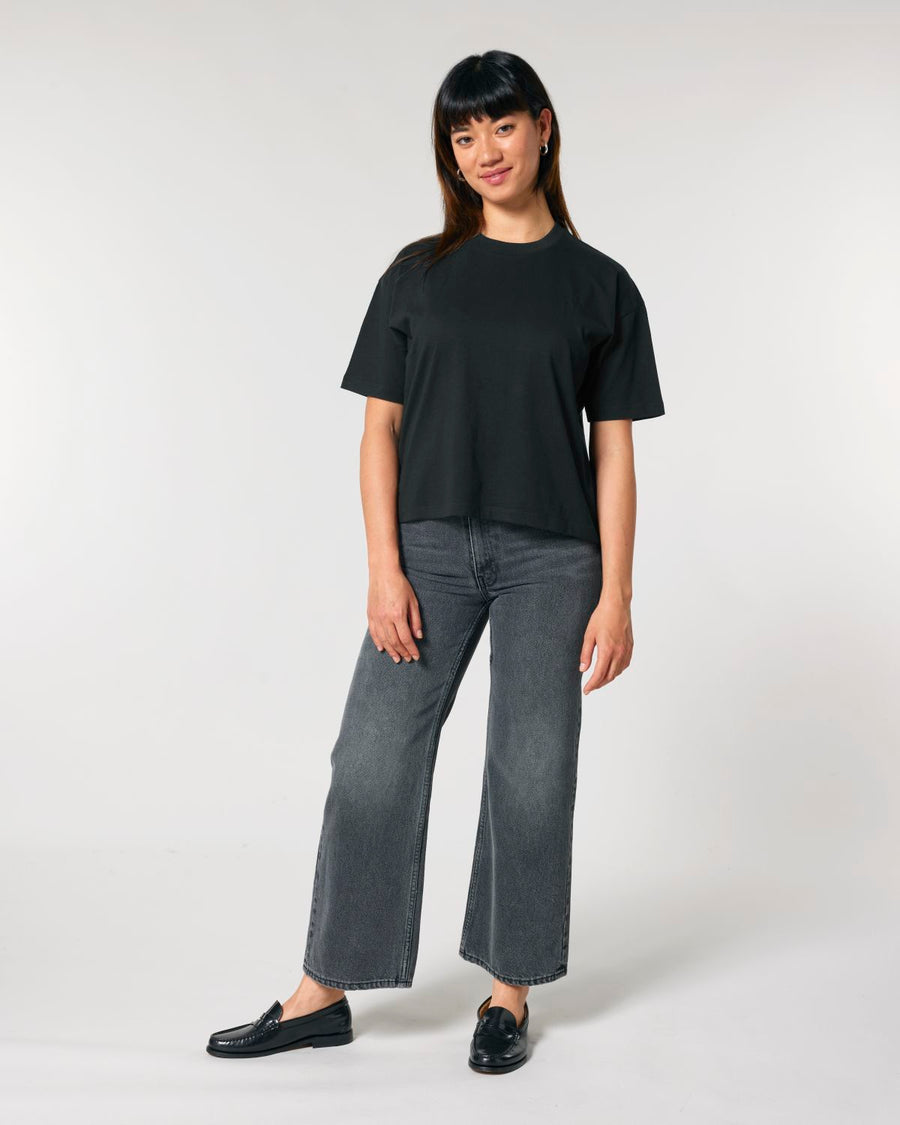 A woman standing against a neutral background, wearing a black Stanley/Stella Women's Boxy Fit T-Shirt made of organic ring-spun cotton, gray jeans, and black loafers.