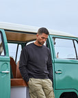 A man in a MyNeedsAreSimple Tee Jays Urban Sweatshirt 5150 Organic Cotton (Label Free) and khaki pants stands with hands in pockets by an open door of a vintage green van.