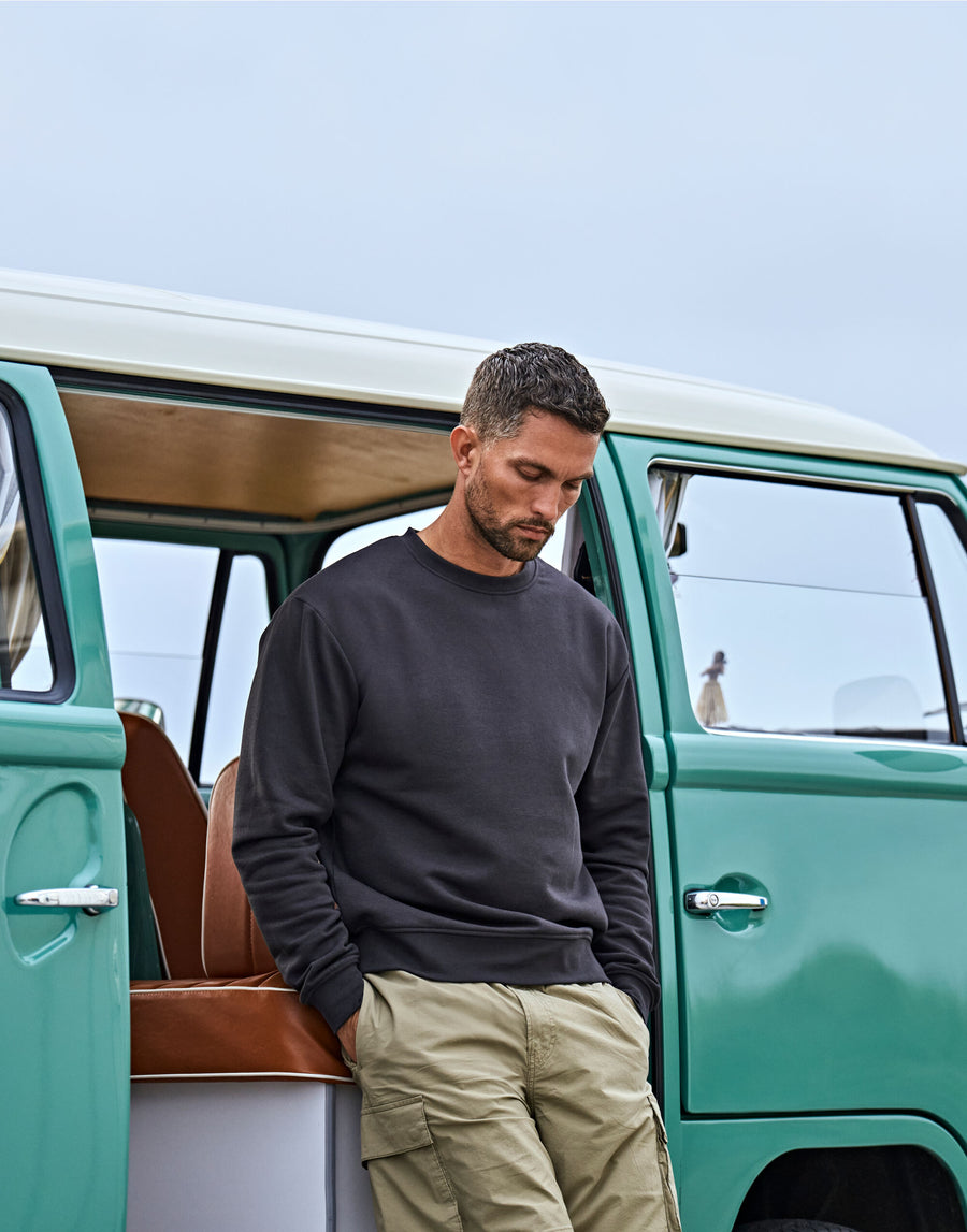 A man in a MyNeedsAreSimple Tee Jays Urban Sweatshirt 5150 Organic Cotton (Label Free) and khaki pants stands with hands in pockets by an open door of a vintage green van.