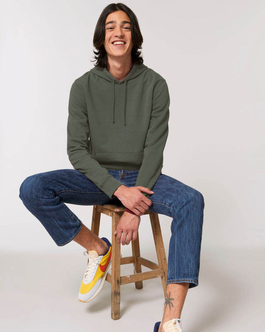 A man sitting on a stool wearing a Stanley/Stella Drummer Hoodie Khaki (C223) and jeans.