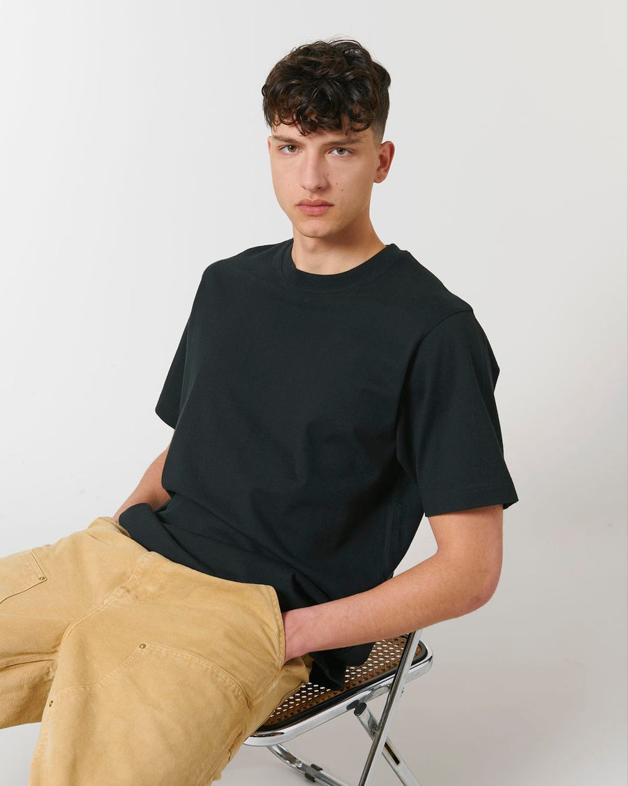 A man sitting on a chair wearing a Stanley/Stella Freestyler Heavy Organic Cotton Unisex T-shirt in fabric washed black and khaki pants.
