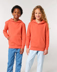 Two children wearing STSK180 Stella/Stella Mini Cruiser 2.0 The Iconic organic cotton kids' hoodie sweatshirts in orange with jeans.