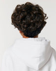 The man is wearing a STSK180 Stella Mini Cruiser 2.0 The Iconic Kids Hoodie Sweatshirt made of organic cotton, with curly hair visible from the back.