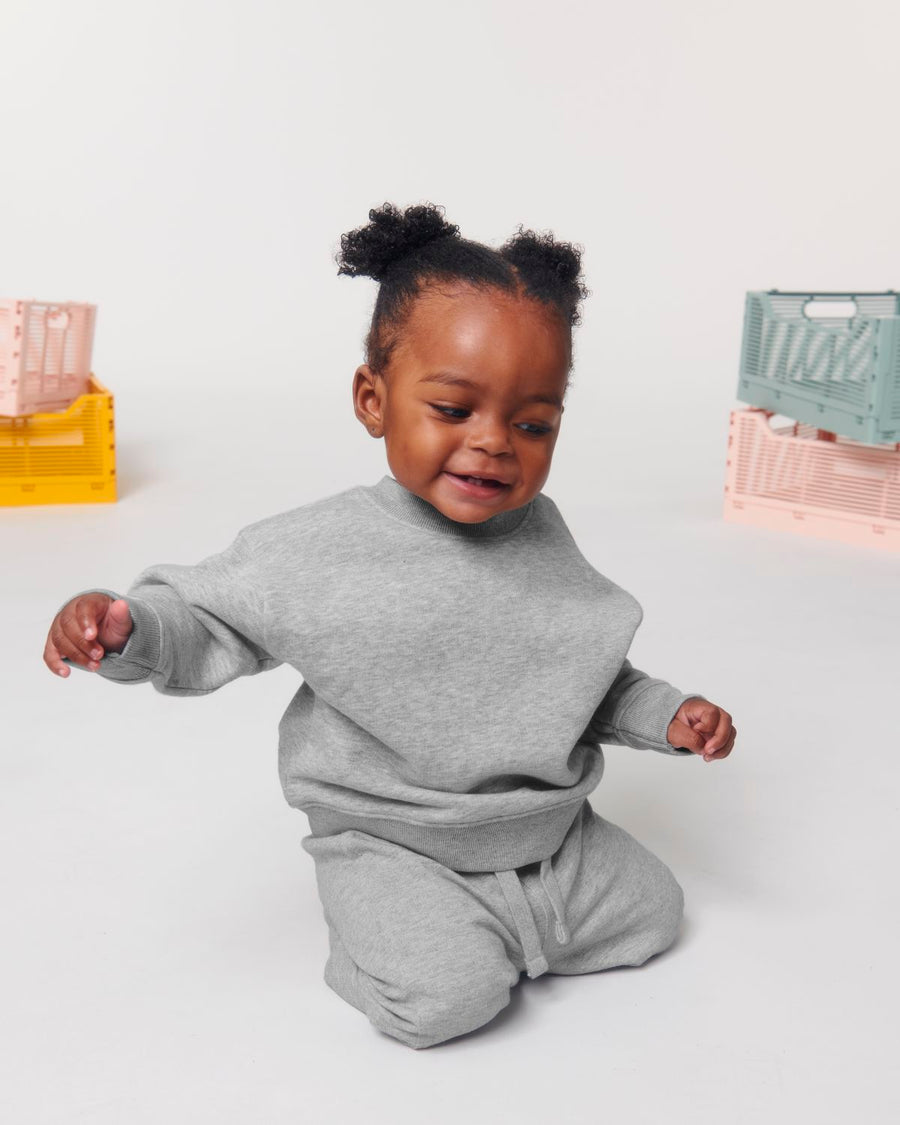 A baby sitting on the floor in a grey sweatsuit made from organic ring-spun cotton STSB920 Stanley/Stella Baby Changer The Iconic Babies' Crew Neck Sweatshirt.