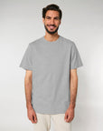 A man with a beard and dark hair is standing against a plain background, wearing a Stanley/Stella STTU171 Stanley/Stella Sparker 2.0 The Unisex Heavy T-Shirt made from 215 GSM Organic Carded Cotton and beige pants, smiling at the camera.