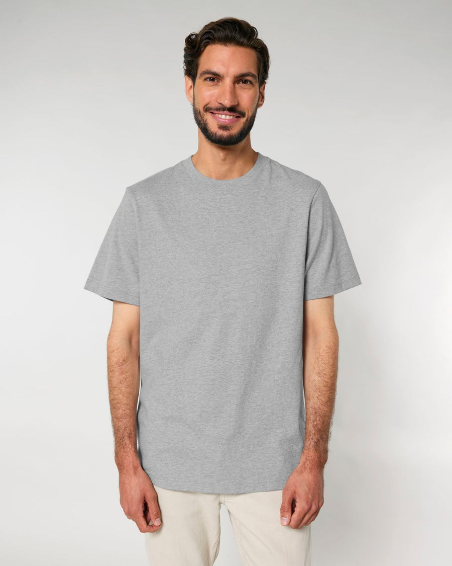 A man with a beard and dark hair is standing against a plain background, wearing a Stanley/Stella STTU171 Stanley/Stella Sparker 2.0 The Unisex Heavy T-Shirt made from 215 GSM Organic Carded Cotton and beige pants, smiling at the camera.