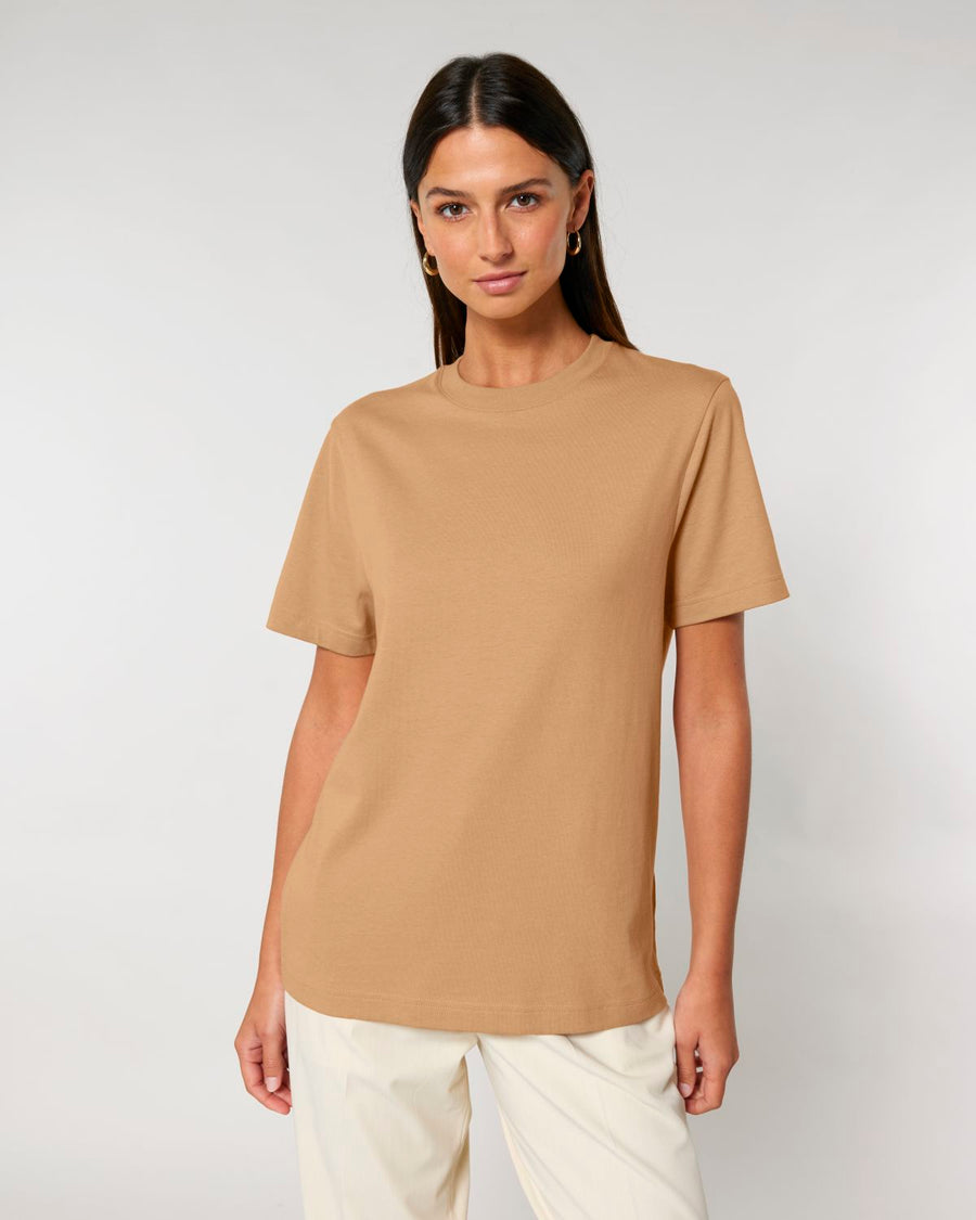 A woman with long dark hair is wearing a plain beige STTU171 Stanley/Stella Sparker 2.0 The Unisex Heavy T-Shirt made of Organic Carded Cotton and white pants, standing against a light gray background.