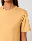 A person wearing a plain, light brown STTU171 Stanley/Stella Sparker 2.0 The Unisex Heavy T-Shirt made from 215 GSM organic carded cotton, shown from the shoulders up against a neutral background. Their face is not visible.