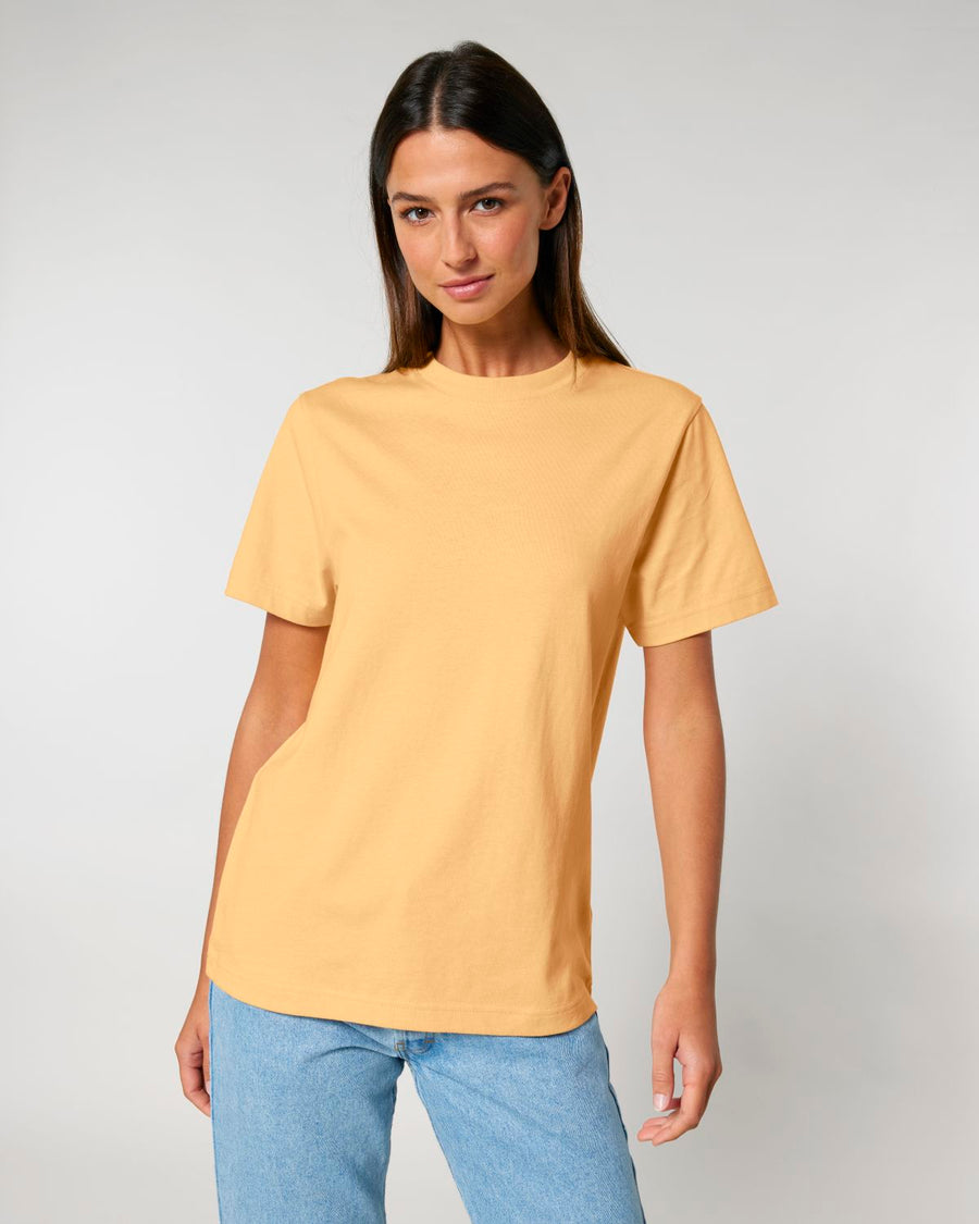 A woman with long brown hair is wearing a plain yellow STTU171 Stanley/Stella Sparker 2.0 The Unisex Heavy T-Shirt made from organic carded cotton and light blue jeans, standing against a plain light gray background.