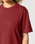 Person wearing a plain, maroon STTU171 Stanley/Stella Sparker 2.0 The Unisex Heavy T-Shirt made from organic carded cotton, with part of a tattoo visible on their forearm.