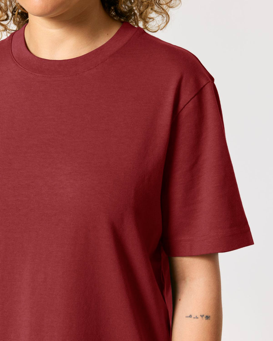 Person wearing a plain, maroon STTU171 Stanley/Stella Sparker 2.0 The Unisex Heavy T-Shirt made from organic carded cotton, with part of a tattoo visible on their forearm.