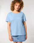 A woman with curly hair is wearing a Stanley/Stella STTW173 Stella Serena Mid-Light Scoop Neck T-Shirt made from 100% Organic Cotton and a denim skirt, standing against a plain background.