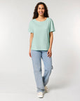 A person with long, curly hair, wearing an STTW173 Stella Serena Mid-Light Scoop Neck T-Shirt by Stanley/Stella made from 100% Organic Cotton in light green and blue jeans, stands against a plain, light background, smiling and facing the camera.