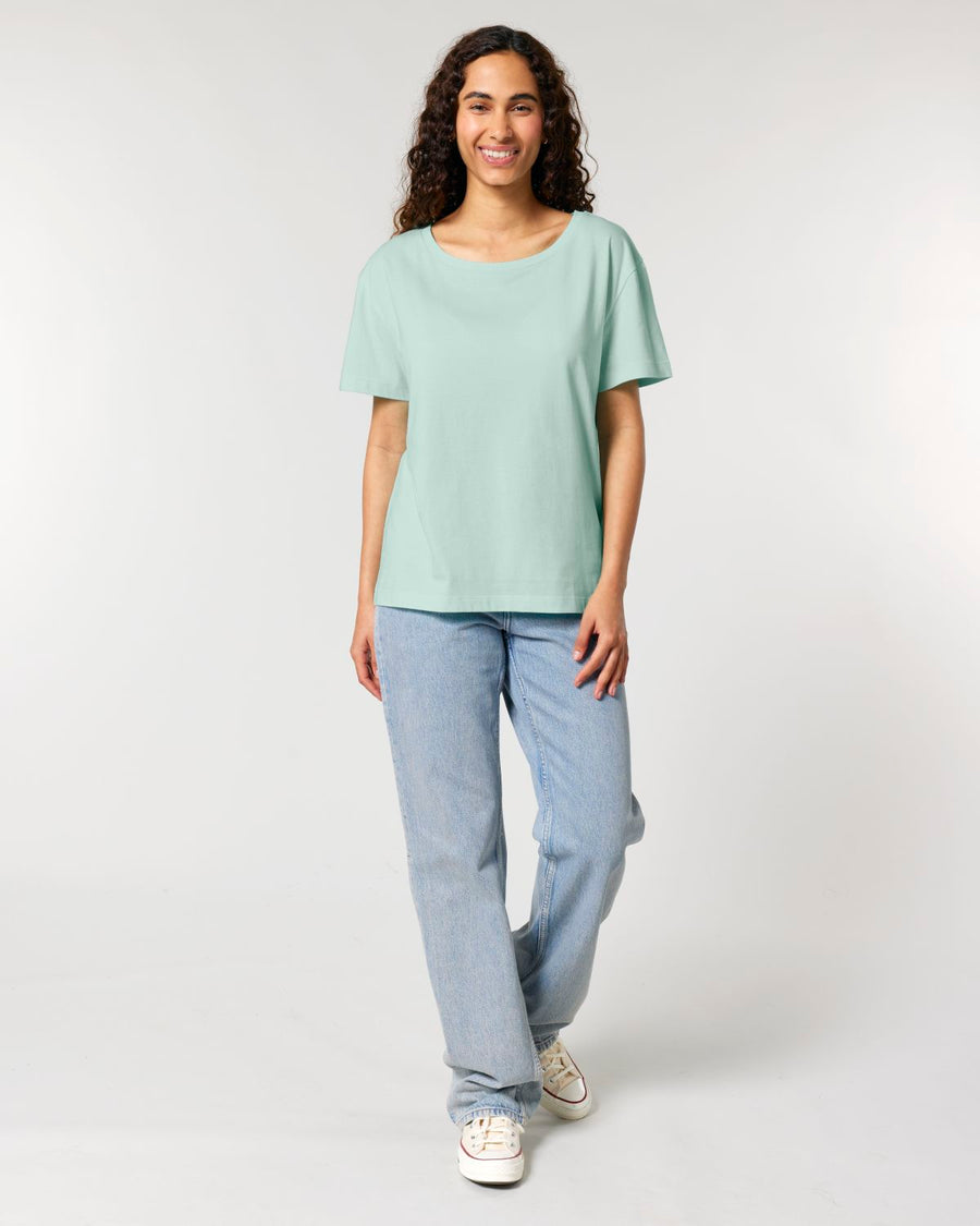 A person with long, curly hair, wearing an STTW173 Stella Serena Mid-Light Scoop Neck T-Shirt by Stanley/Stella made from 100% Organic Cotton in light green and blue jeans, stands against a plain, light background, smiling and facing the camera.