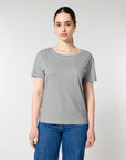 A person with long hair pulled back is wearing a Stanley/Stella STTW173 Stella Serena Mid-Light Scoop Neck T-Shirt in 100% Organic Cotton and blue jeans, standing against a plain white background.