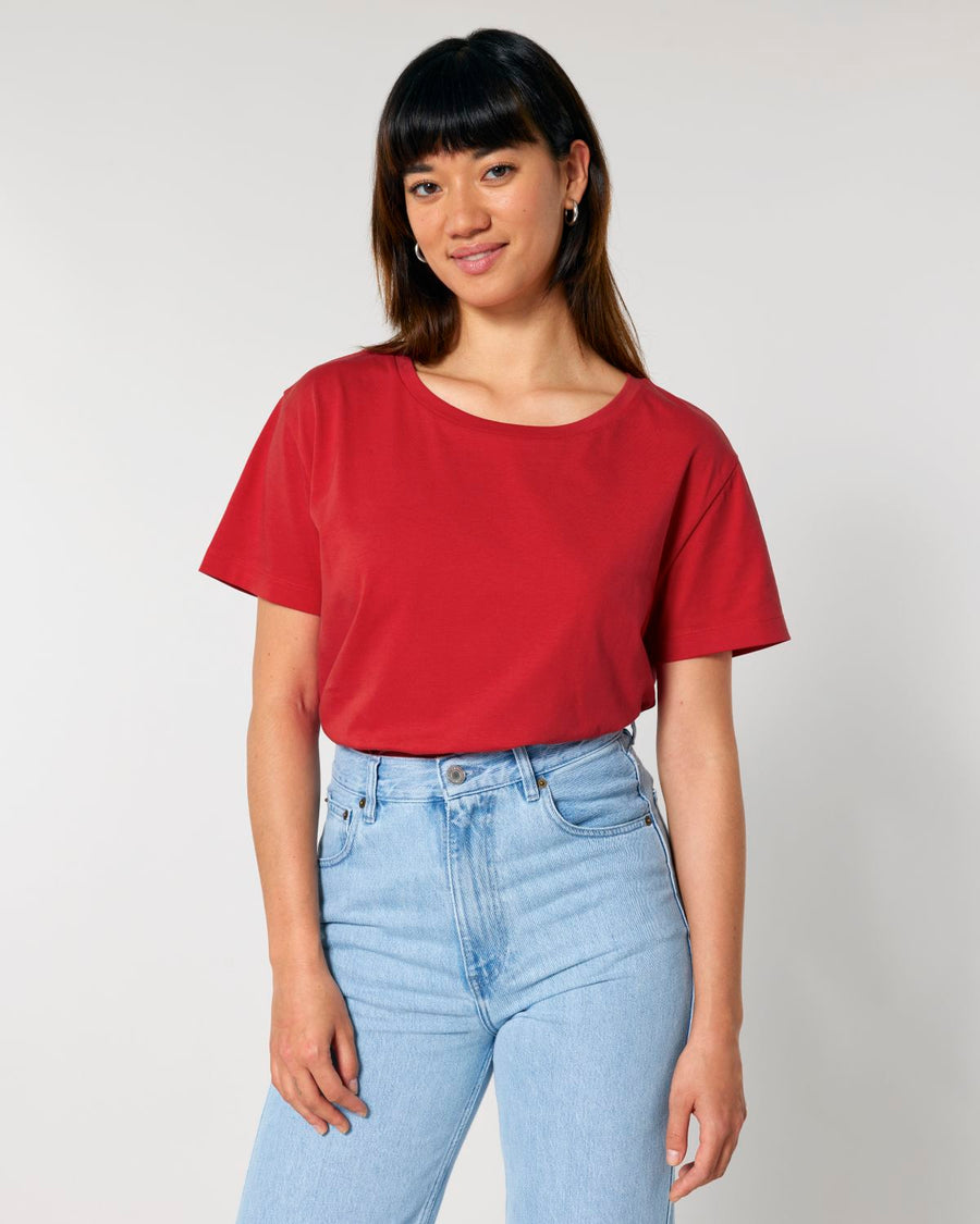 A person with long dark hair wearing an iconic Stanley/Stella STTW173 Stella Serena Mid-Light Scoop Neck T-Shirt in red, made of 100% organic cotton, paired with light blue jeans standing against a plain background.