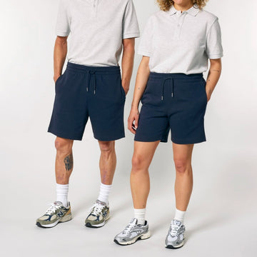 Two people stand side by side wearing matching grey shirts, Stanley/Stella STBU186 Stella/Stella Trainer 2.0 The Iconic Mid-light Unisex Jogger Shorts in navy blue, white socks, and athletic shoes.